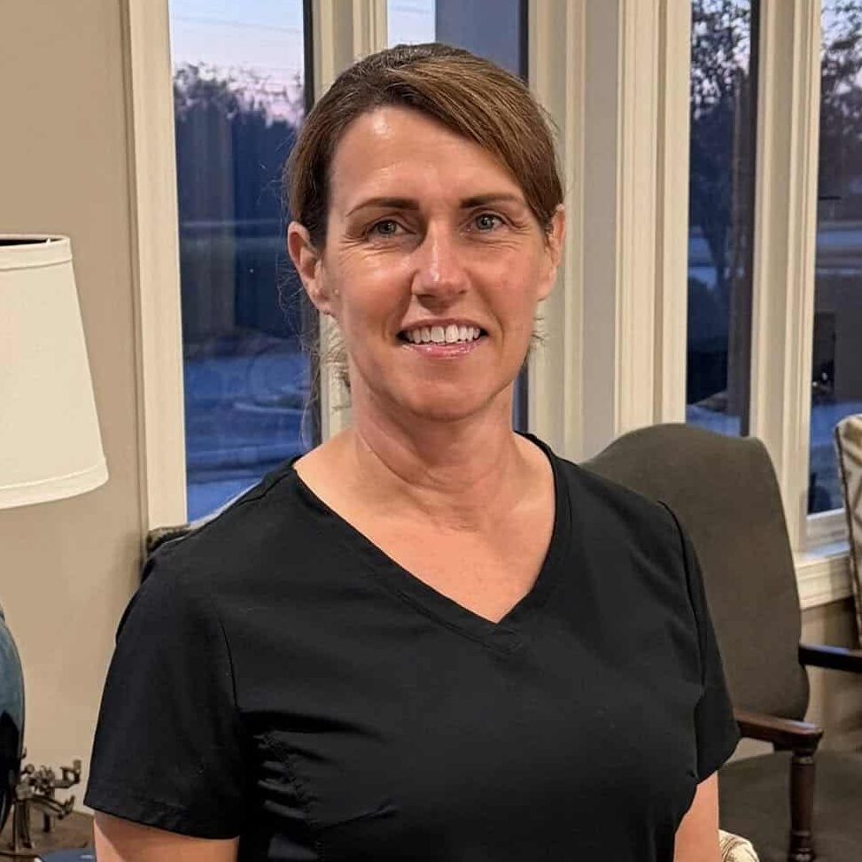 A woman in black scrubs smiles in a warmly lit office or waiting area with big windows, chairs, a lamp, and framed airplane art.