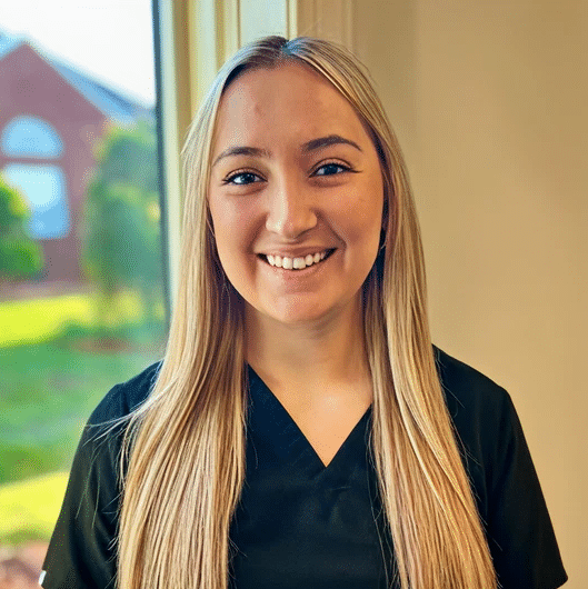 A young woman with long blonde hair in black scrubs smiles indoors by a window, with a green lawn and brick building outside.