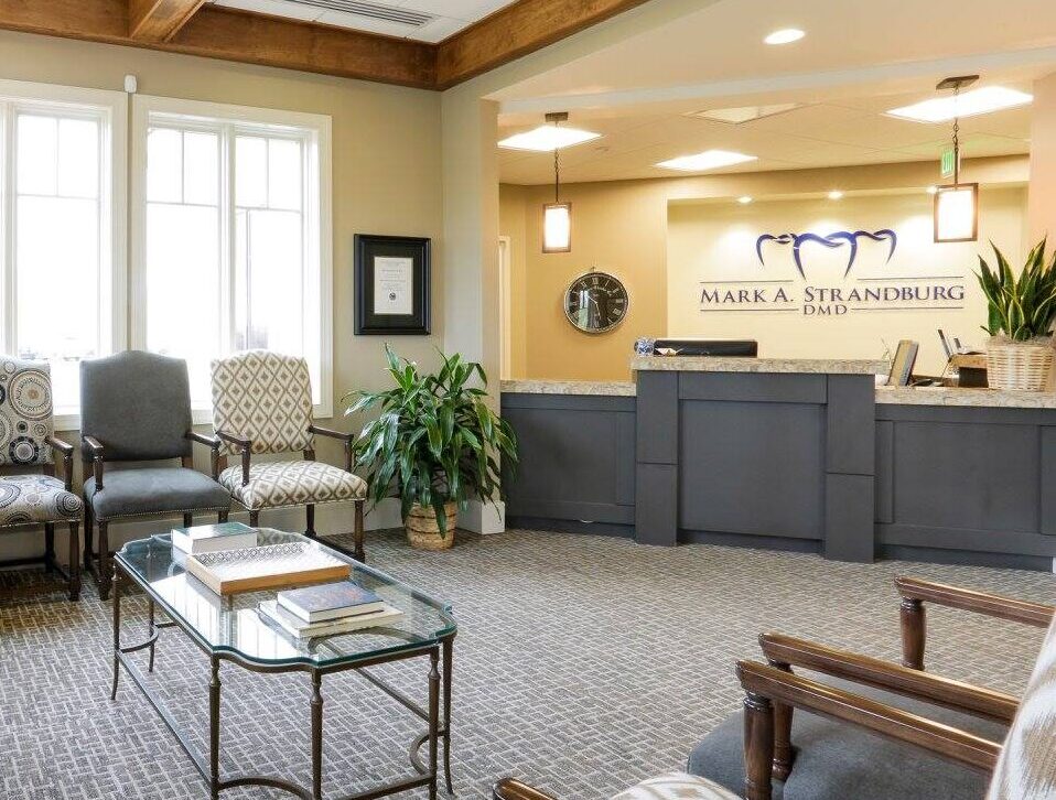 A bright, welcoming waiting room with patterned chairs, a glass coffee table, potted plants, and a reception desk in the background. Large windows let in natural light.