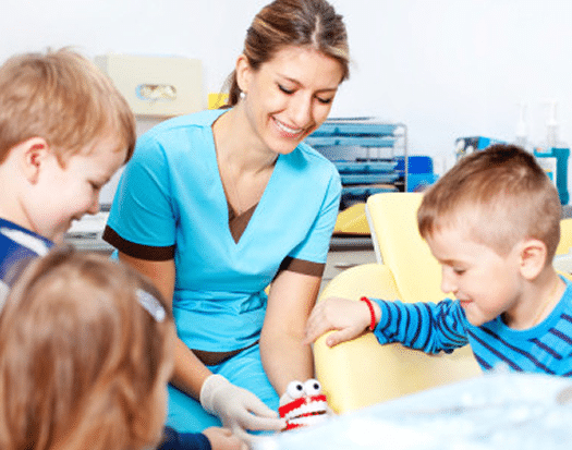 A woman in medical scrubs smiles as she shows dental tools to three engaged young children gathered closely around her.