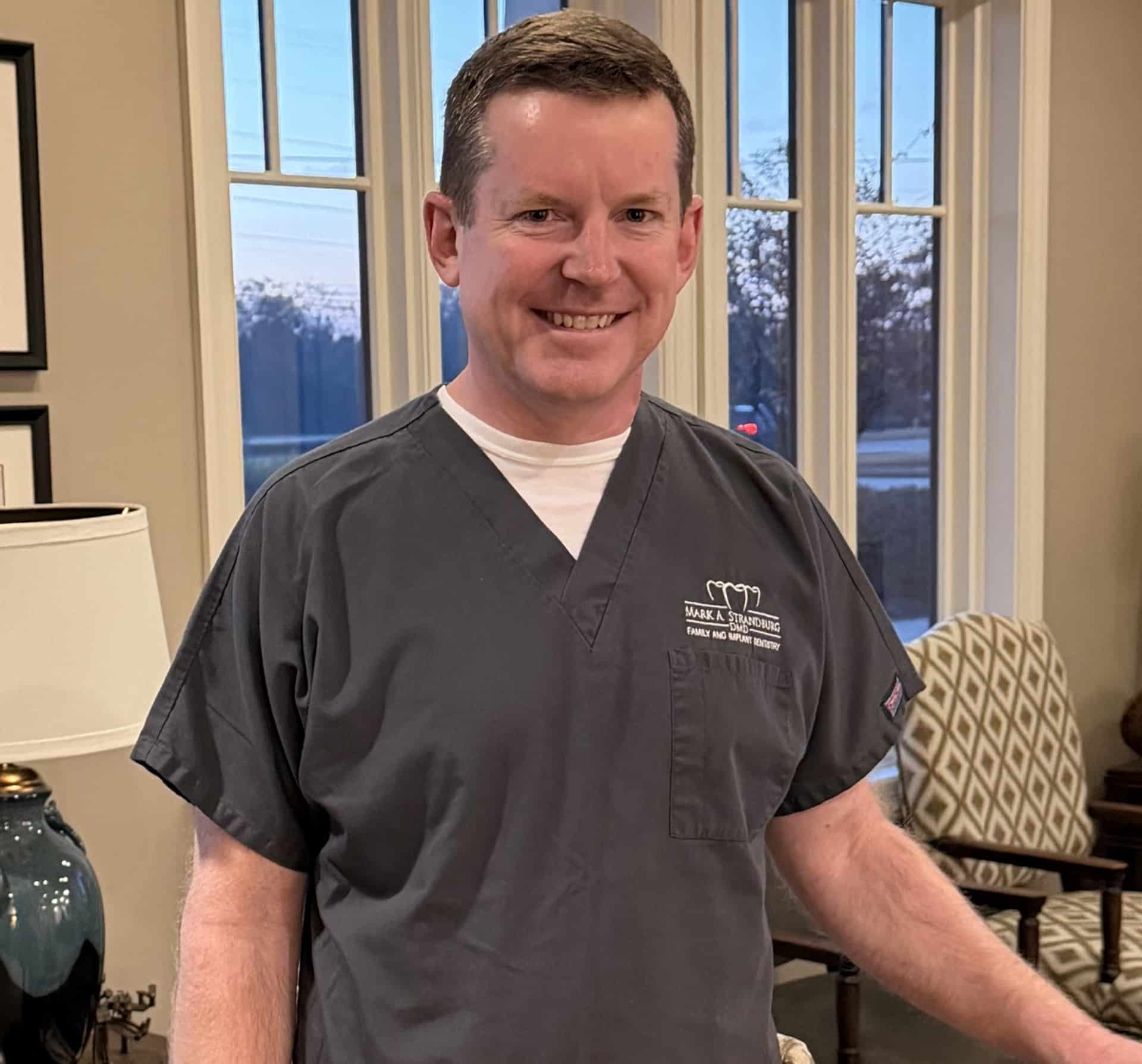 A man in gray medical scrubs stands indoors, smiling at the camera with large windows, an armchair, and a table with a lamp behind him.
