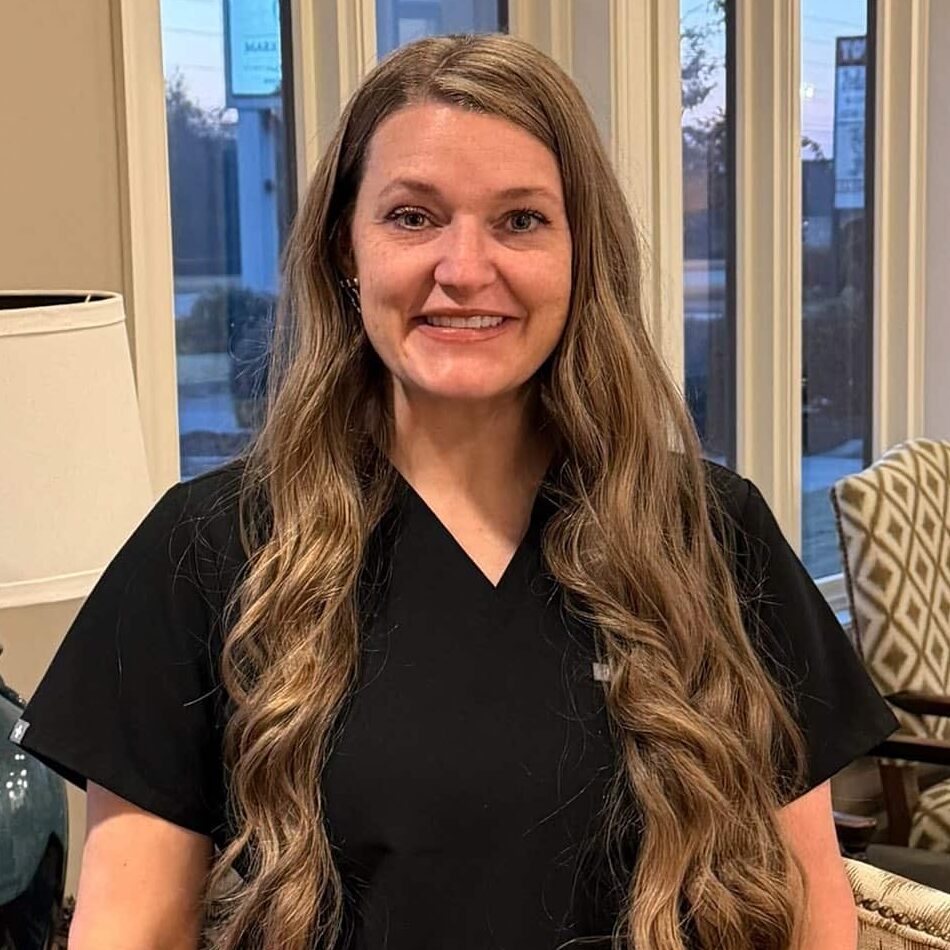 A woman with long wavy hair in black scrubs smiles in a bright room with big windows, armchairs, lamps, and aircraft art.