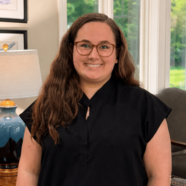A woman with long brown hair and glasses smiles in a living room by a blue lamp, airplane art, and big windows with green outside.