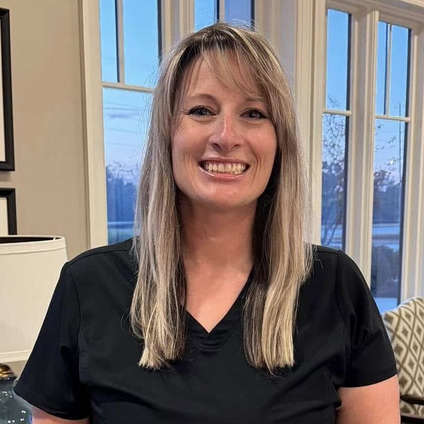 A smiling blonde woman in black scrubs stands in a bright office with wooden furniture, artwork, and large windows in the background.