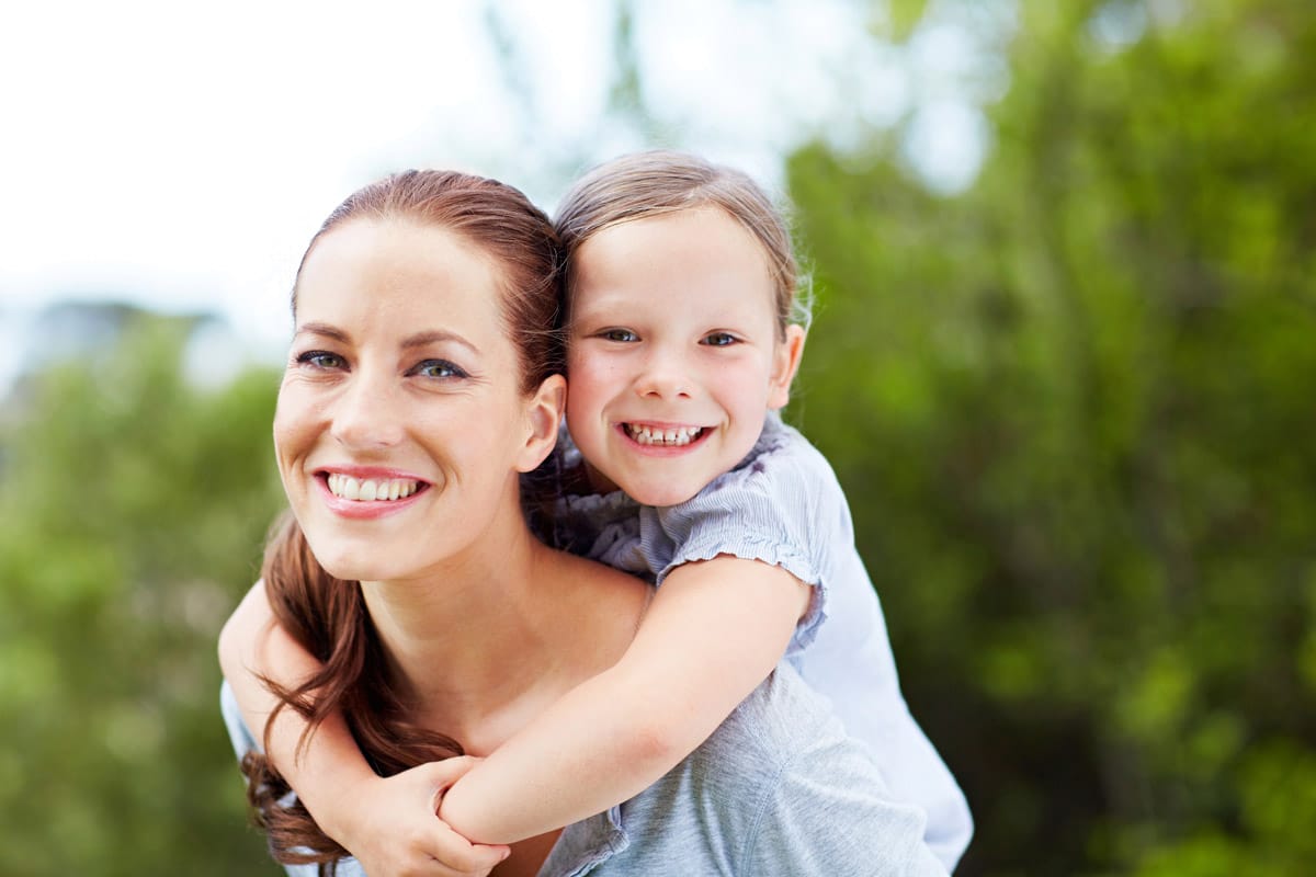 A smiling woman with brown hair gives a piggyback ride to a young girl, both looking at the camera outside with greenery behind them.