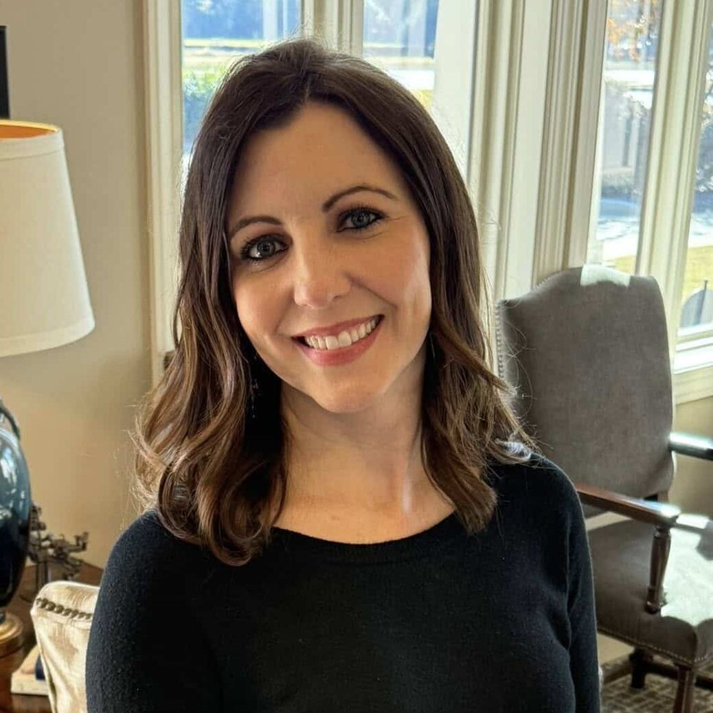 A woman with shoulder-length brown hair in a black sweater and plaid pants smiles in a cozy room with large windows and patterned chairs.